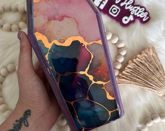 May include: Coffin-shaped tumbler with a vibrant abstract design. The tumbler features a mix of pink, blue, and gold colors. The text "Tumbler Hustler" is visible on a purple background.