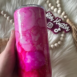 May include: A pink and magenta tumbler with a watercolor effect and gold glitter accents. The stainless steel tumbler has a clear lid and is held by a hand. The tumbler has the text "Tumbler Hustler" on a purple tag.