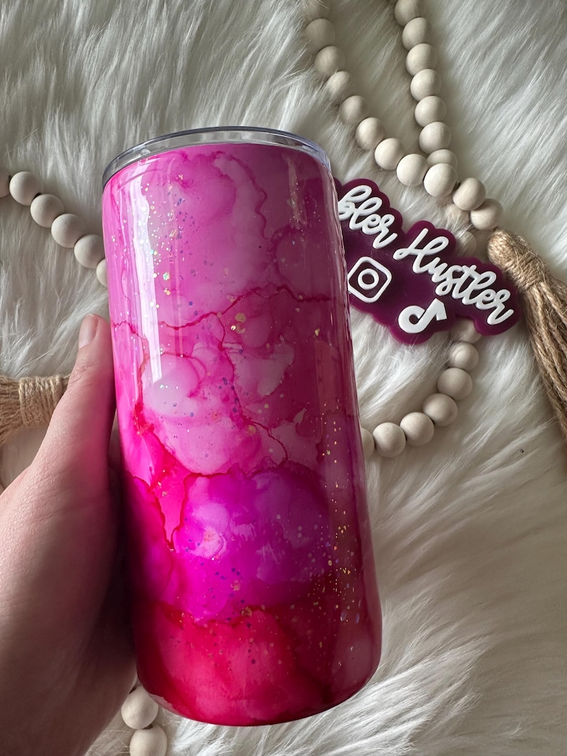 May include: A pink and magenta tumbler with a watercolor effect and gold glitter accents. The stainless steel tumbler has a clear lid and is held by a hand. The tumbler has the text "Tumbler Hustler" on a purple tag.