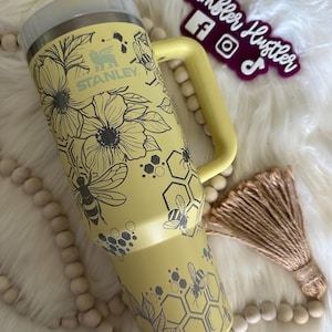 Könnte beinhalten: Gelber Stanley-Becher mit Blumen- und Bienenmuster. Der Becher hat einen Griff, einen Strohhalm und einen Deckel. Das Design umfasst Blumen, Bienen und Wabenmuster. Das Stanley-Logo ist sichtbar.