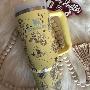 Könnte beinhalten: Ein gelber Stanley-Becher mit Griff, Strohhalm und Deckel. Der Becher ist mit schwarzen Illustrationen im Harry-Potter-Stil verziert. Der Becher hat das Stanley-Logo und den Text "Hogwarts Express".