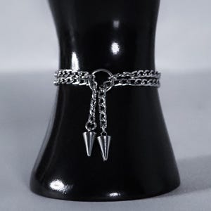 May include: A silver-toned metal chain bracelet featuring two delicate chains connected by a central ring. Two short chain segments hang from the ring, each ending in a small, pointed, cone-shaped metal charm. The bracelet has a polished finish.