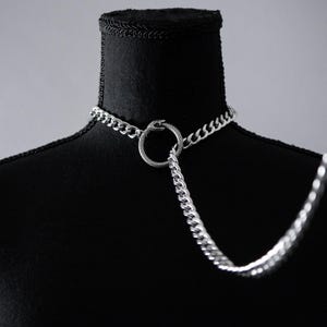 May include: Silver-toned chain choker necklace with a snake-shaped ring pendant. The necklace is displayed on a black mannequin, with a long chain extending downwards. The jewelry is likely made of stainless steel.