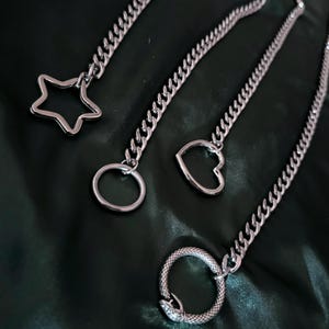May include: Three silver chains with different charms: a star, a heart, and a snake. The chains are on a dark green fabric background.