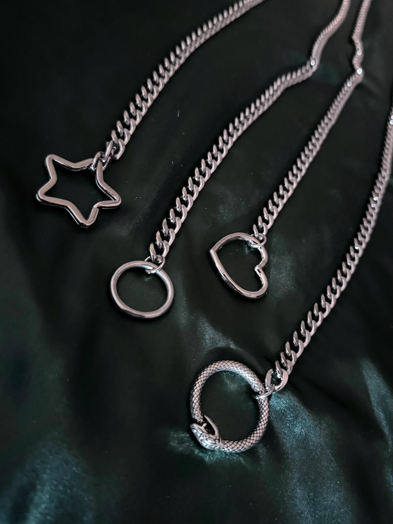 May include: Three silver chains with different charms: a star, a heart, and a snake. The chains are on a dark green fabric background.