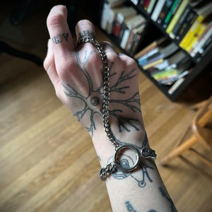 May include: A silver chain bracelet with a large ring. The bracelet is on a person's wrist, which is tattooed with a neuron and chemical structures.