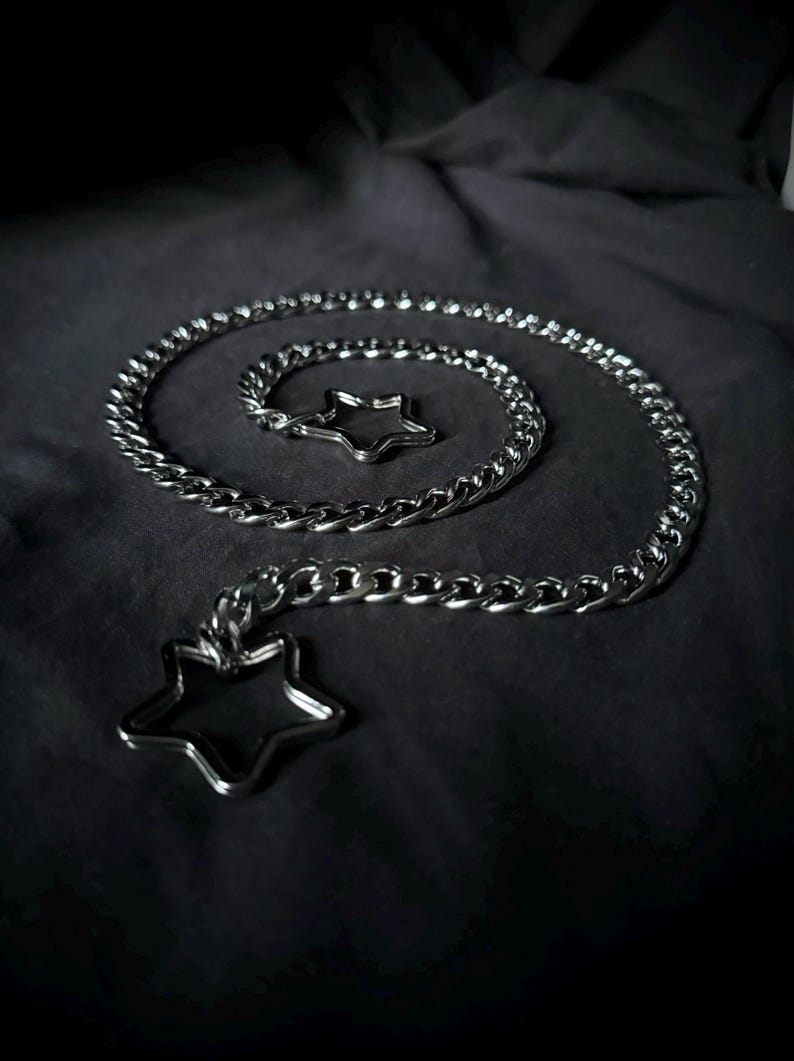 May include: A silver chain necklace with two star-shaped charms. The chain is thick and has a curb link design. The charms are simple and elegant.