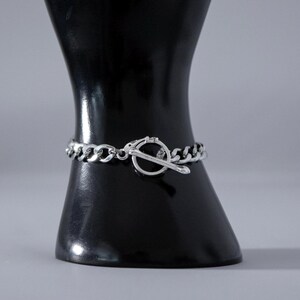 May include: A silver chain bracelet with a toggle clasp. The bracelet is displayed on a black mannequin. The chain links are thick and rounded. The toggle clasp is a circular design with a bar that slides through it to secure the bracelet.
