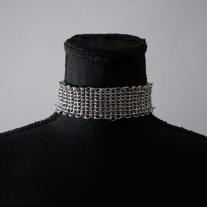 May include: A silver chainmail choker necklace displayed on a black mannequin. The choker is made of small, interlocking metal rings, creating a woven texture. The necklace sits around the mannequin's neck, showcasing its design.