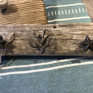 May include: A rustic wooden wall hook with three metal hooks shaped like a starfish, a coral branch, and another starfish. The wood is weathered and gray, and the hooks are a dark bronze color. The text "Local Drift" is written on a small tag attached to the hook.