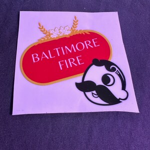 May include: A sticker featuring the text "BALTIMORE FIRE" in white on a red oval with a gold border. Above the text are gold wheat and maple leaf designs. A cartoon character with a mustache is on the right.