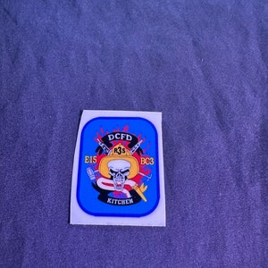 May include: A blue and white sticker with a skull and crossbones design. The text "DCFD" is at the top of the sticker, and the text "R3S" is below the skull. The text "E15" is on the left side of the skull, and the text "BC3" is on the right side of the skull. The text "KITCHEN" is at the bottom of the sticker.