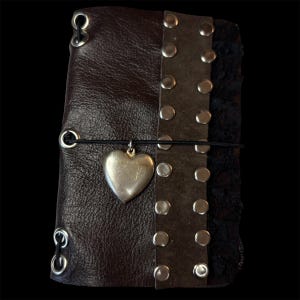 May include: A dark brown leather journal with a silver heart charm. The journal has a riveted leather strap and a black elastic closure. The edges are lined with black fabric and silver eyelets.
