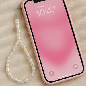 Könnte beinhalten: Ein pinkfarbenes Smartphone in einer pinkfarbenen Hülle mit einer Perlen-Handschlaufe. Der Bildschirm zeigt einen rosa Farbverlaufshintergrund mit der Uhrzeit 10:47 und den Worten "Swipe up to unlock".