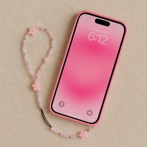 Könnte beinhalten: Ein pinkfarbenes Smartphone in einer pinkfarbenen Hülle mit einer Perlenkette. Der Bildschirm zeigt die Uhrzeit 6:12 Uhr am Dienstag, 9. April. Die Kette hat rosa Blumen- und Perlenakzente.