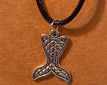 May include: A silver mermaid tail pendant necklace on a black cord, displayed on a brown card with a hole at the top. The pendant has a detailed scale pattern.