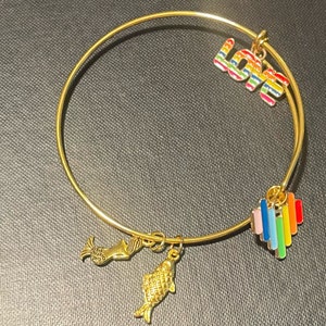Puede incluir: Un brazalete de oro con abalorios de colores arcoíris. Los abalorios incluyen la palabra "LOVE", un corazón, una sirena y un pez.