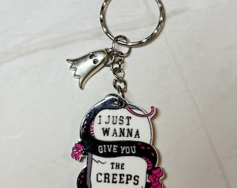 Puede incluir: Un llavero plateado con un dije de fantasma y un colgante rectangular. El colgante presenta una serpiente negra enrollada alrededor de un letrero blanco con el texto "I JUST WANNA GIVE YOU THE CREEPS" y flores rosas.
