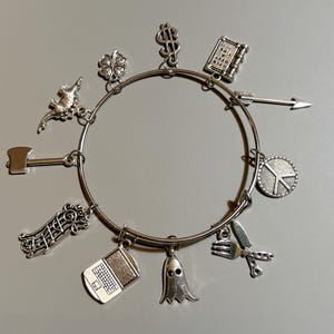 Op de afbeelding: Zilveren bedelarmband met diverse bedels, waaronder een laptop, een spook, een vredesteken en een pijl. Andere bedels zijn een klavertjevier, een dollarteken en een kleine bijl. De armband wordt getoond op een lichtgrijze ondergrond.