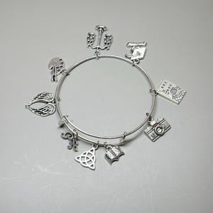 May include: Silver-toned charm bracelet featuring various charms, including an anchor, a camera, a book, a Celtic knot, and angel wings. The bracelet is expandable and has a simple, elegant design.