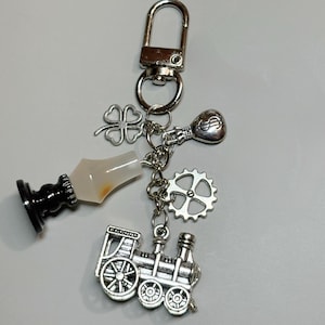 Op de afbeelding: Zilverkleurige sleutelhanger met diverse bedels. Bedels zijn onder meer een klavertjevier, een gloeilamp, een tandwiel en een miniatuurtrein. De sleutelhanger heeft ook een zwart-witte flesstopper.