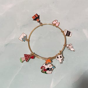 May include: Gold-tone charm bracelet featuring various enamel charms. Charms include a skull with mushrooms, a coffee cup, a ghost, a cat, a mushroom, a four-leaf clover, and an envelope. The bracelet is a stylish accessory.