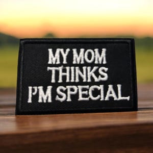 May include: Black rectangular patch with white embroidered text that reads "MY MOM THINKS I'M SPECIAL". The patch is likely made of fabric and is displayed on a wooden surface with a blurred background.