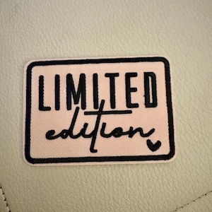 May include: A rectangular patch with a pink background and a black border. The words "LIMITED edition" are printed on the patch in black, with the word "edition" in a cursive font. A small black heart is at the bottom.