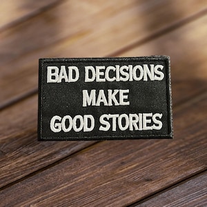 May include: Black fabric patch with white embroidered text that reads "Bad Decisions Make Good Stories".
