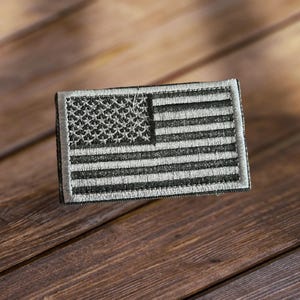 May include: A small, embroidered American flag patch in black and white. The patch is on a wooden surface.
