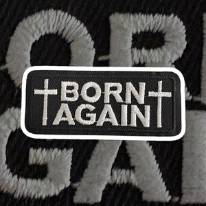May include: A black patch with white embroidered text that reads "BORN AGAIN" with a cross on each side. The patch has a white border and is set against a black background with the embroidered letters "OR" and "GA" in white.