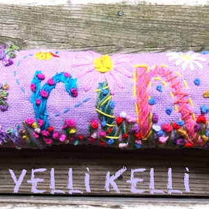 May include: A lavender rectangular pillow embroidered with the name "YELLI KELLI" in colorful thread. The letters are decorated with flowers, vines, and a bird. The pillow is a decorative item.