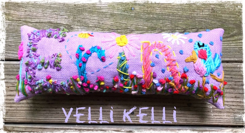 May include: A lavender rectangular pillow embroidered with the name "YELLI KELLI" in colorful thread. The letters are decorated with flowers, vines, and a bird. The pillow is a decorative item.