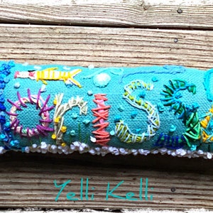 May include: A turquoise blue pillow with the name "CRISS" embroidered in colorful thread. The letters are surrounded by various sea creatures and plants, including a seahorse, a fish, and seaweed. The pillow has a white trim around the edges.
