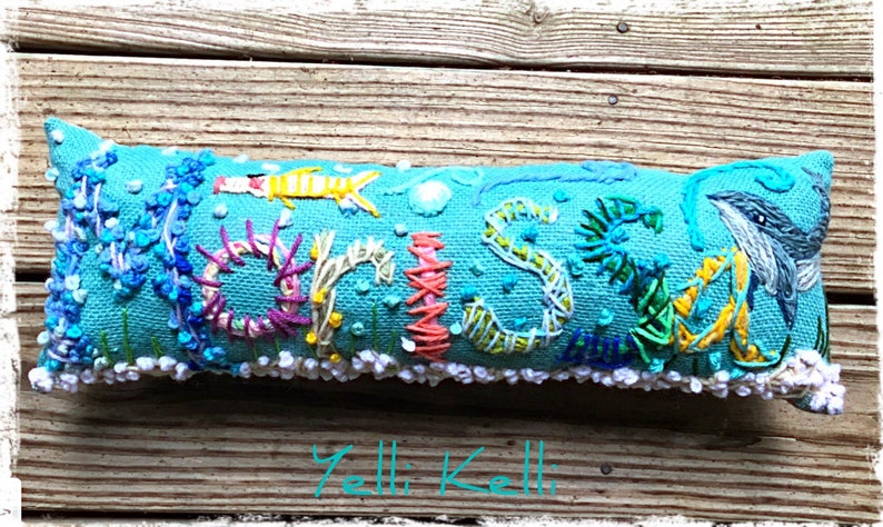 May include: A turquoise blue pillow with the name "CRISS" embroidered in colorful thread. The letters are surrounded by various sea creatures and plants, including a seahorse, a fish, and seaweed. The pillow has a white trim around the edges.