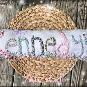 May include: A white pillow with the name "Kennedy" embroidered in colorful thread. The pillow is decorated with beads and a small flower.
