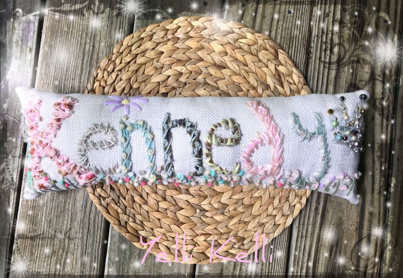 May include: A white pillow with the name "Kennedy" embroidered in colorful thread. The pillow is decorated with beads and a small flower.