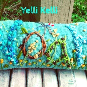 May include: A rectangular, turquoise pillow with embroidered floral and animal designs. The word "MOM" is embroidered in the center, with a deer on the left and a white flower on the right. The text "Yelli Kelli" is at the top.