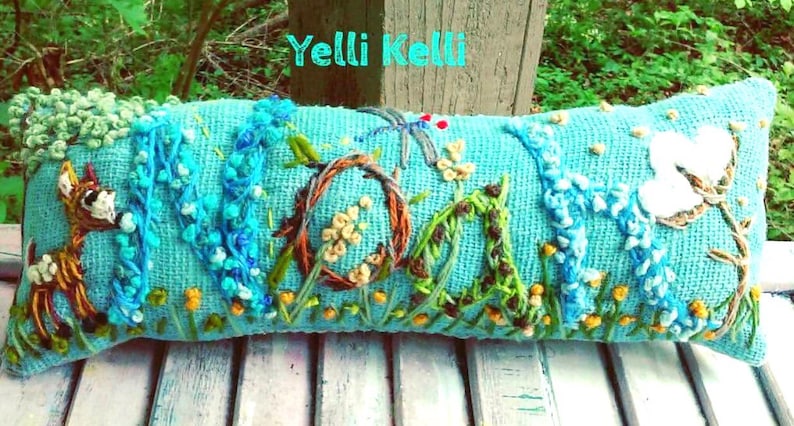 May include: A rectangular, turquoise pillow with embroidered floral and animal designs. The word "MOM" is embroidered in the center, with a deer on the left and a white flower on the right. The text "Yelli Kelli" is at the top.