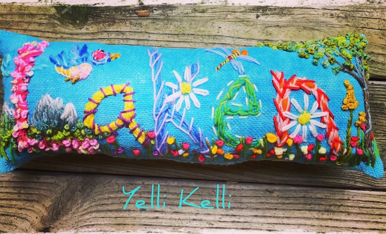 May include: Embroidered blue pillow with the word "LAKE" in colorful thread. The design includes a bird, dragonfly, flowers, and a tree. The text "Yelli Kelli" is at the bottom.