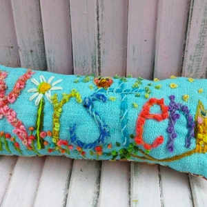 May include: A turquoise blue pillow with the name "Kristen" embroidered in colorful thread. The pillow is decorated with embroidered flowers and a small owl.