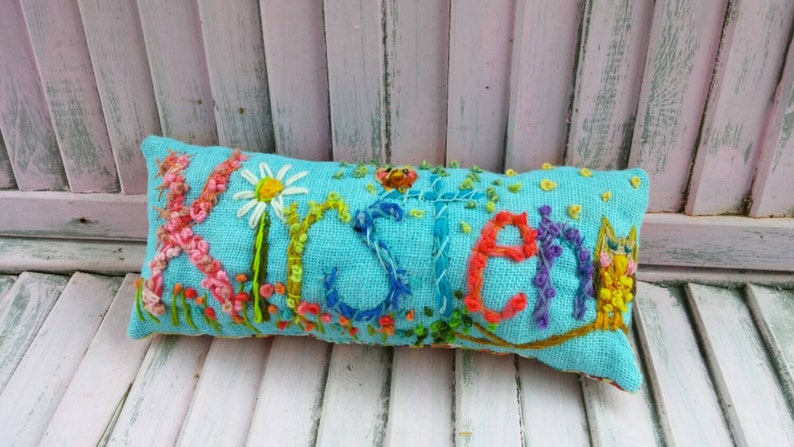 May include: A turquoise blue pillow with the name "Kristen" embroidered in colorful thread. The pillow is decorated with embroidered flowers and a small owl.