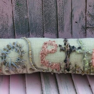 May include: A light green fabric pillow with the word "Celia" embroidered in colorful thread. The pillow is decorated with various textures and patterns, including beads and sequins.