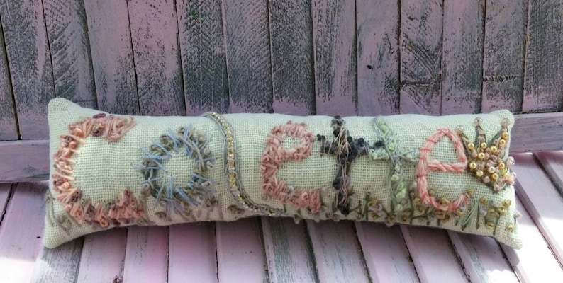 May include: A light green fabric pillow with the word "Celia" embroidered in colorful thread. The pillow is decorated with various textures and patterns, including beads and sequins.