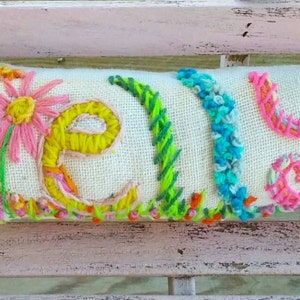 May include: Embroidered rectangular pillow with the name "Kelly" in colorful yarn. The letters are surrounded by embroidered flowers, grass, and a blue bird. The pillow is on a light-colored surface.