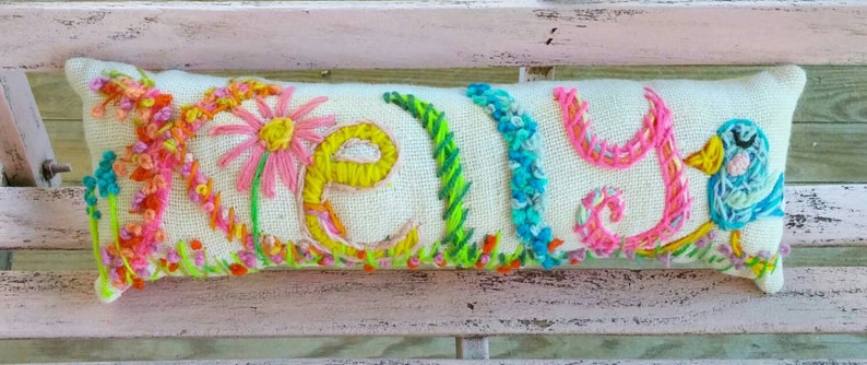 May include: Embroidered rectangular pillow with the name "Kelly" in colorful yarn. The letters are surrounded by embroidered flowers, grass, and a blue bird. The pillow is on a light-colored surface.