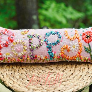 May include: A rectangular, light pink pillow embroidered with the word "POPPY" in colorful thread. The letters are surrounded by embroidered flowers, including daisies and red blooms. The pillow is resting on a woven, circular mat.