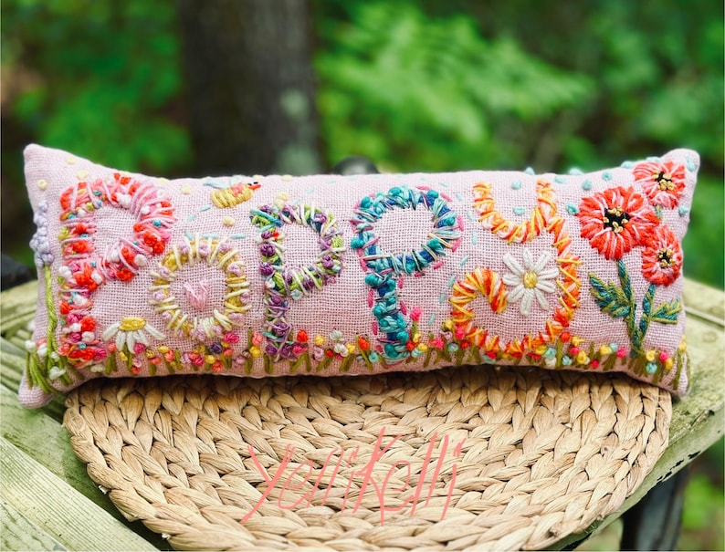 May include: A rectangular, light pink pillow embroidered with the word "POPPY" in colorful thread. The letters are surrounded by embroidered flowers, including daisies and red blooms. The pillow is resting on a woven, circular mat.