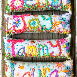 May include: Four embroidered pillow covers with the names "Betty", "Daisy", "Tammy", and "Laura" spelled out in colorful thread. The pillows are white with a textured fabric and feature a variety of colorful flowers and other designs.