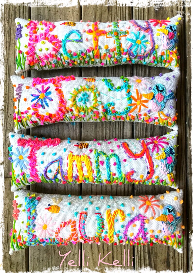 May include: Four embroidered pillow covers with the names "Betty", "Daisy", "Tammy", and "Laura" spelled out in colorful thread. The pillows are white with a textured fabric and feature a variety of colorful flowers and other designs.